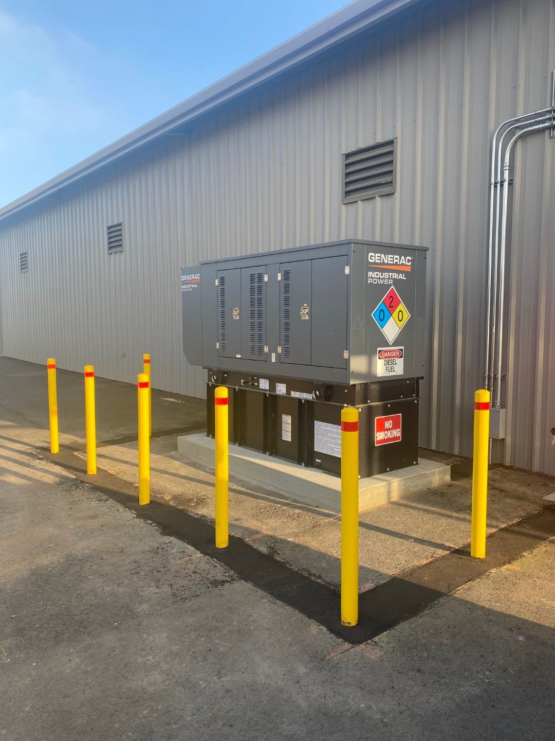 Outdoor electrical equipment beside a gray building, surrounded by yellow safety bollards and hazard labels.