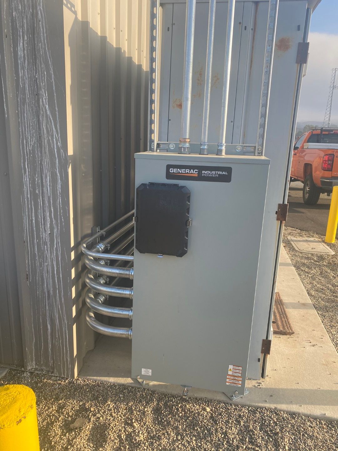 Gray utility box with coiled metal conduit beside a corrugated wall, near a sidewalk and parked vehicle.