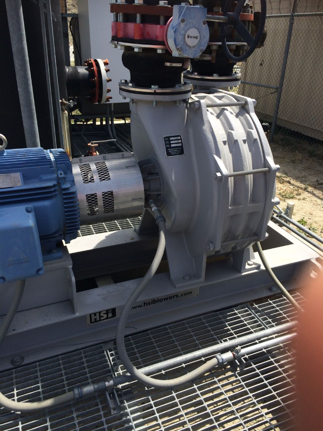 Blue electric motor driving a gray industrial blower on a metal platform with piping and valves