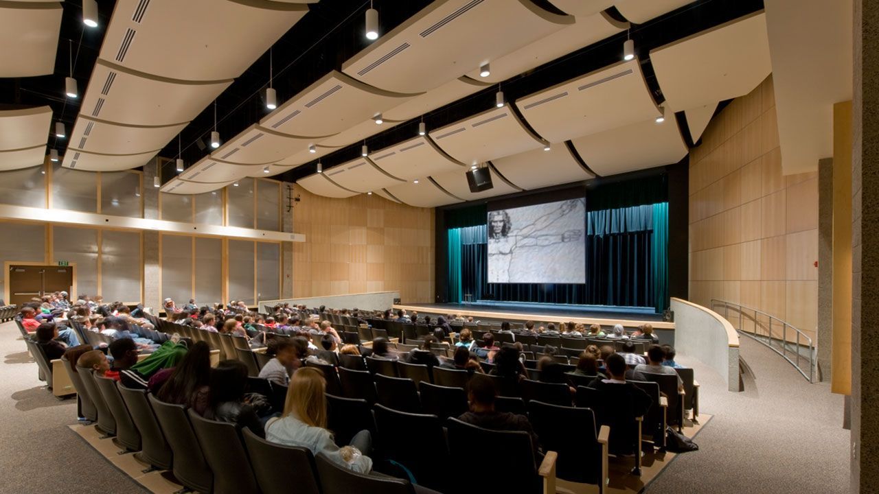 Conference hall filled with seated attendees facing a stage and large projection screen at the front.