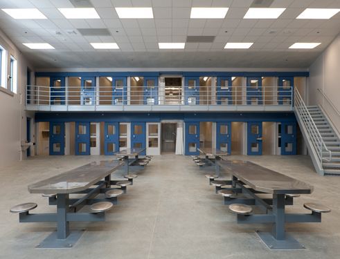 Empty prison common room with blue cell doors, upper catwalk, and metal tables with benches