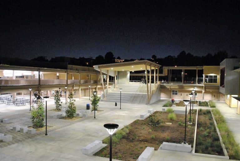Nighttime courtyard with paved walkways, benches, trees, and a lit pavilion between campus buildings