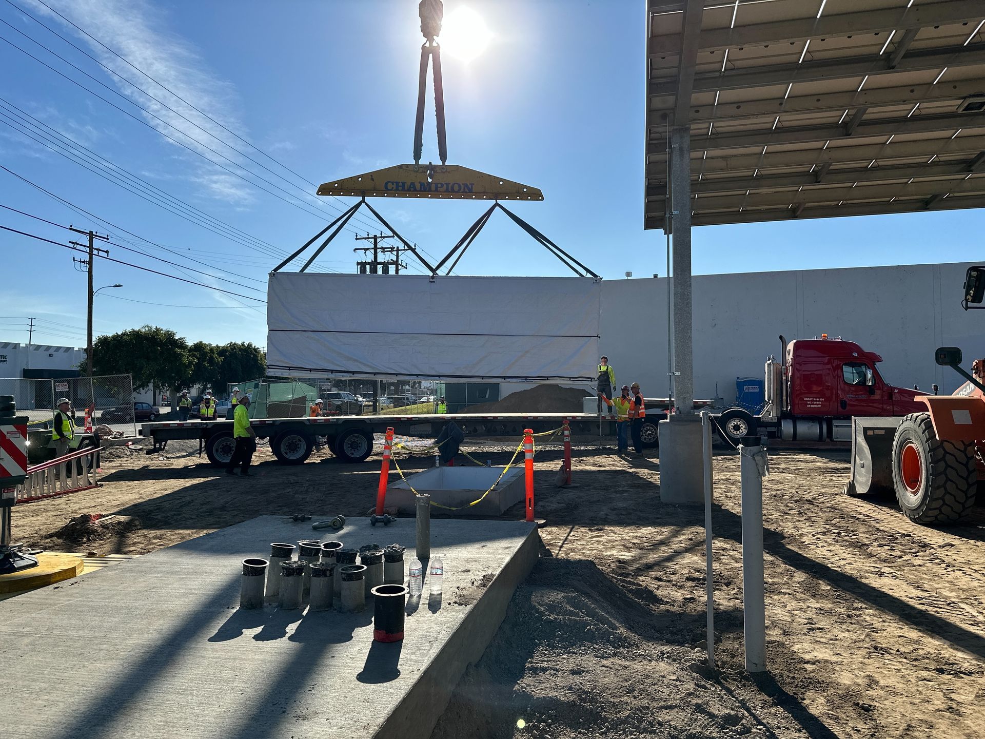Construction site with crane lifting a large white panel near trucks and orange cones under bright sun