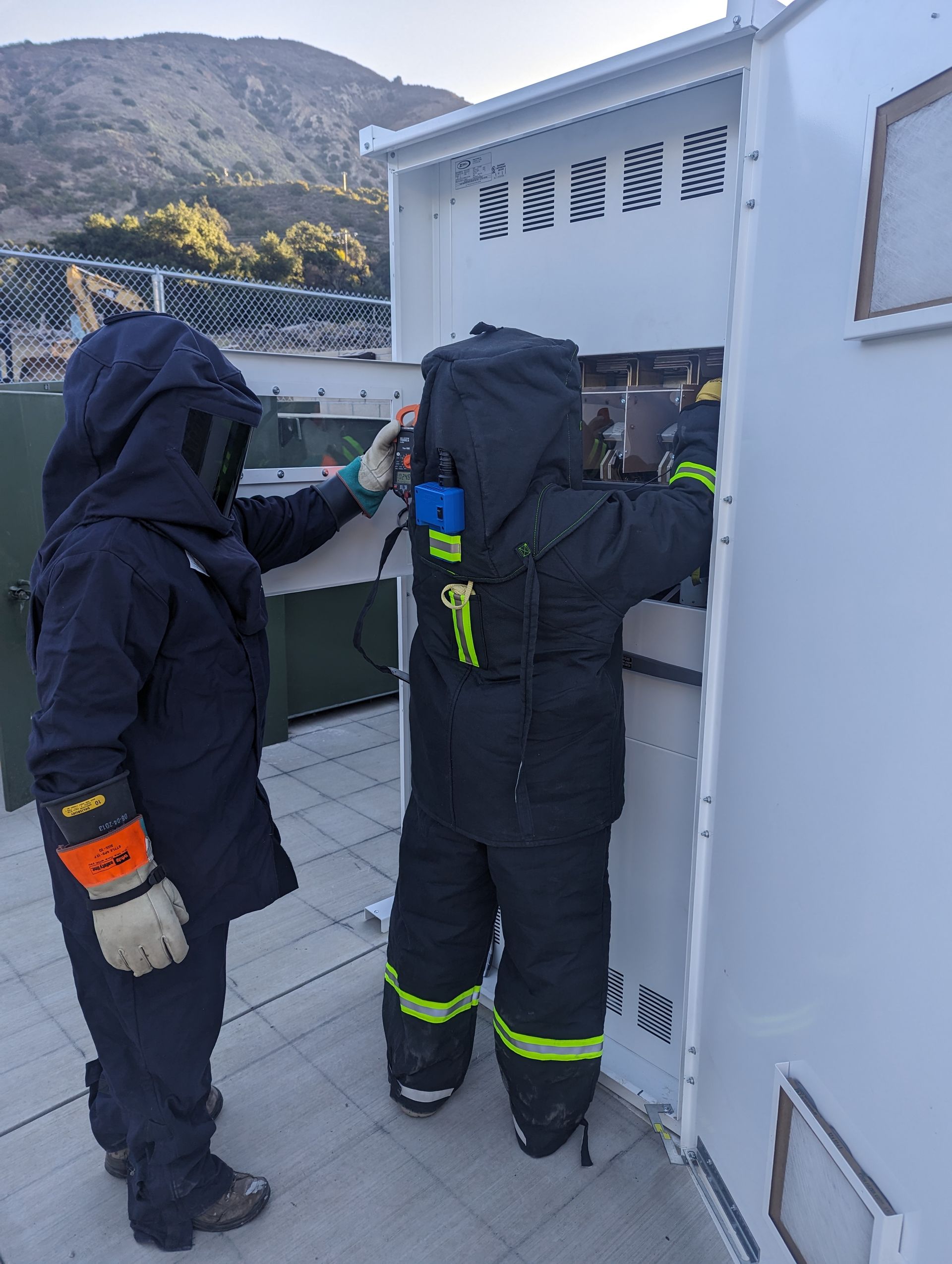 Two people in dark protective suits working at an outdoor utility panel by a building.
