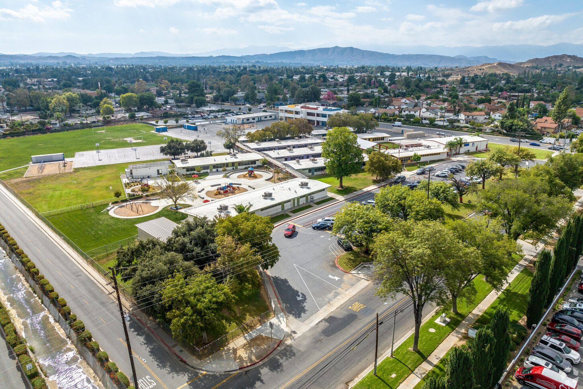 Aerial view of a suburban cityscape with roads, buildings, trees, and distant hills under a clear sky