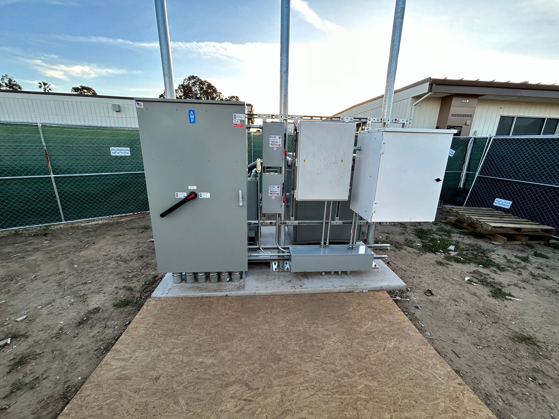 Outdoor electrical utility cabinets and equipment on a concrete pad beside a fenced area.