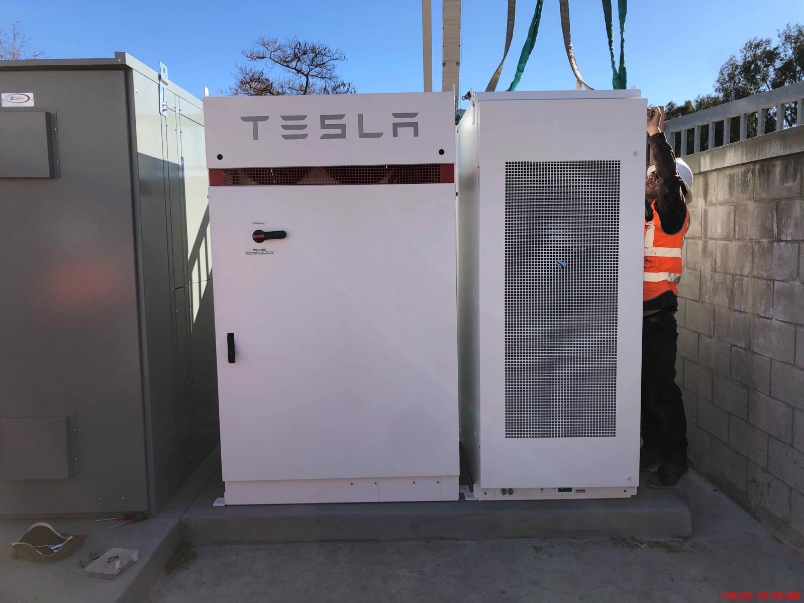 Tesla battery storage units outdoors beside a wall, with a worker in orange safety gear nearby.