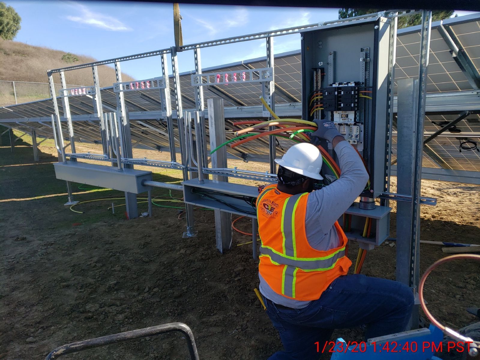 Worker in hard hat and safety vest kneels beside a metal structure, tightening bolts outdoors.