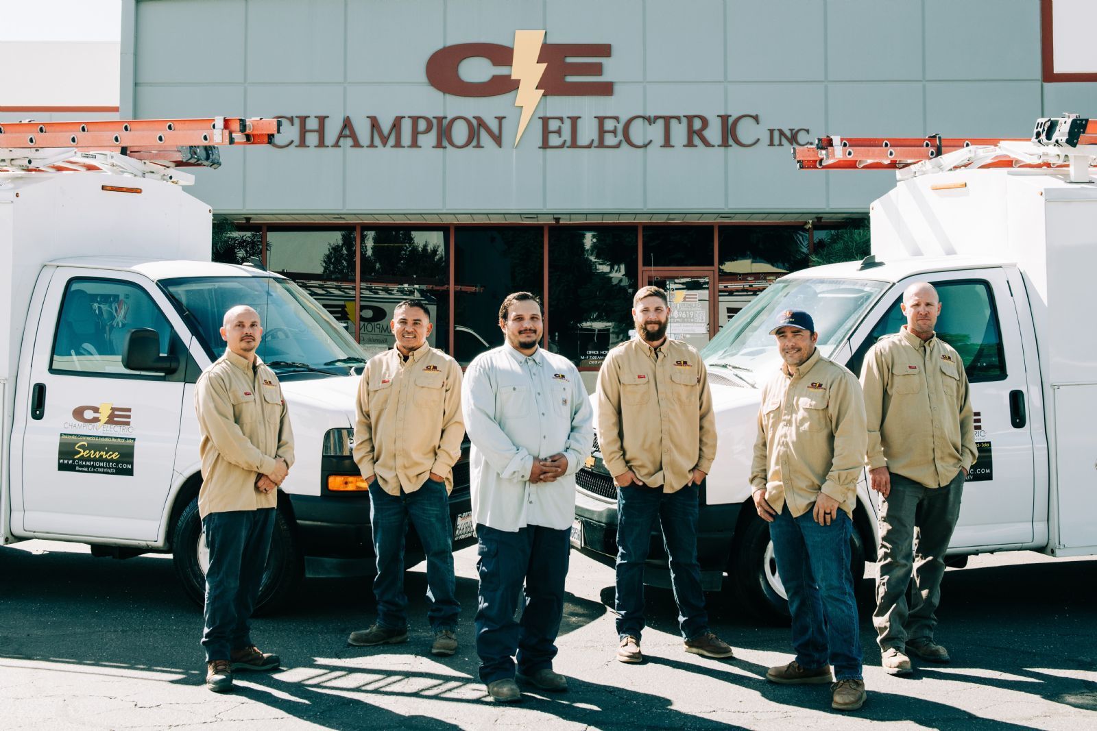 Six workers stand in front of Champion Electric vans at a company building.