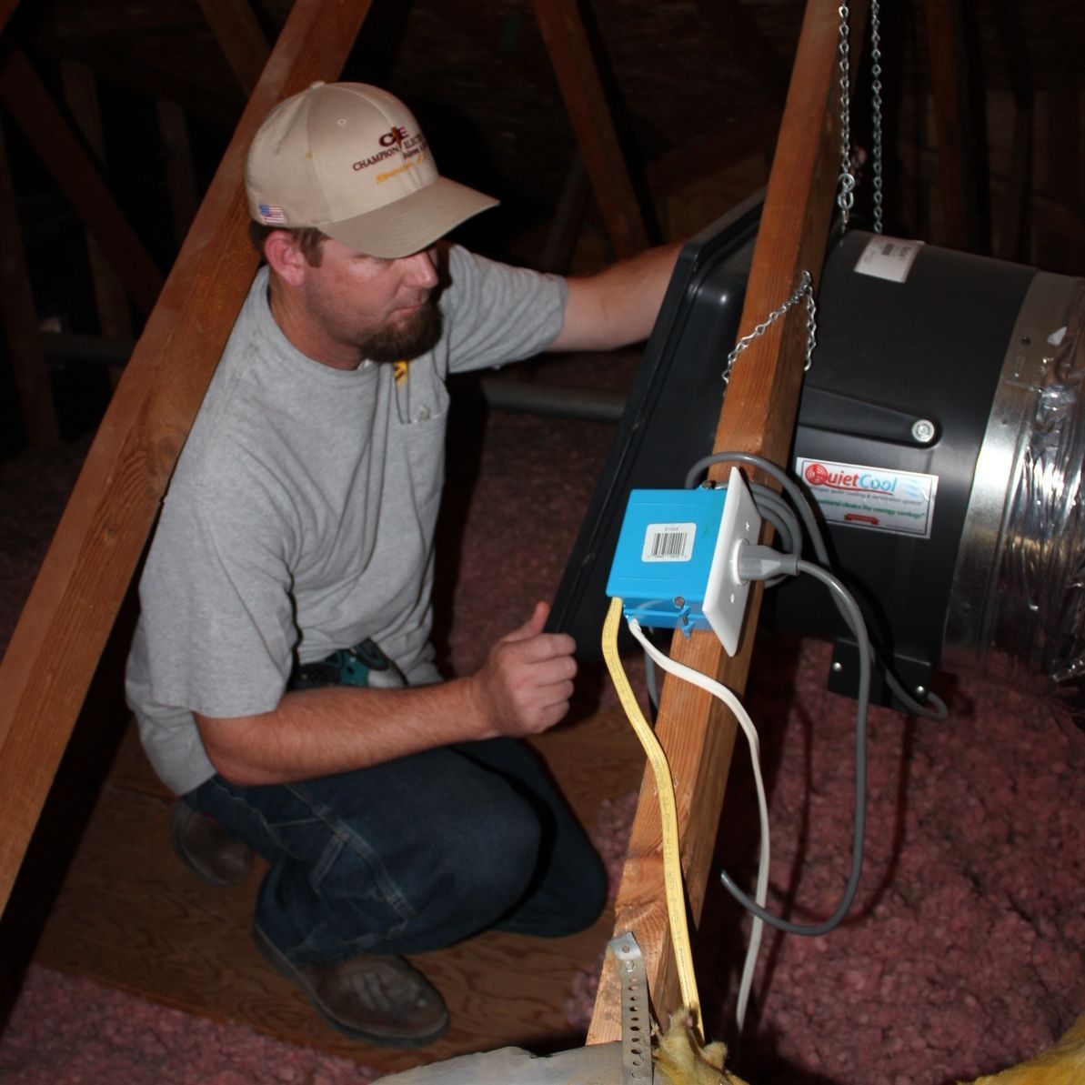 Man kneeling beside a duct fan, adjusting a blue device with cables in a basement space