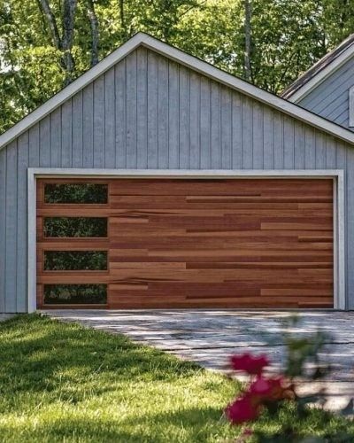 Wooden garage door with horizontal planks, three glass panels, set in a gray-blue building, fronted by green lawn.