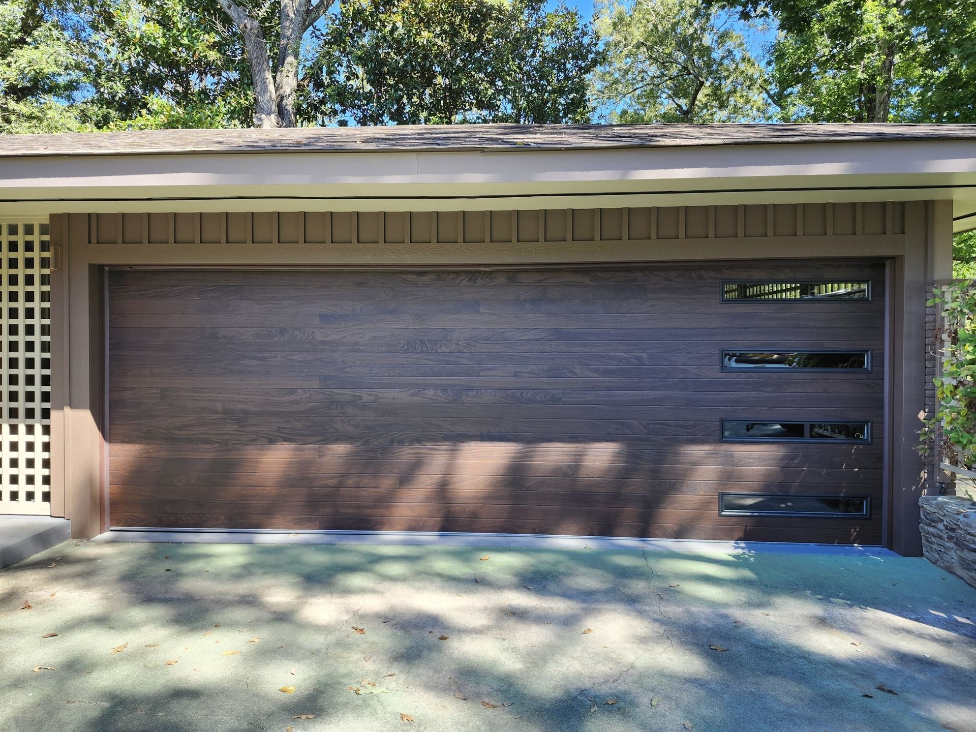 Brown garage door with horizontal glass windows, under a brown roof, in a shaded area.