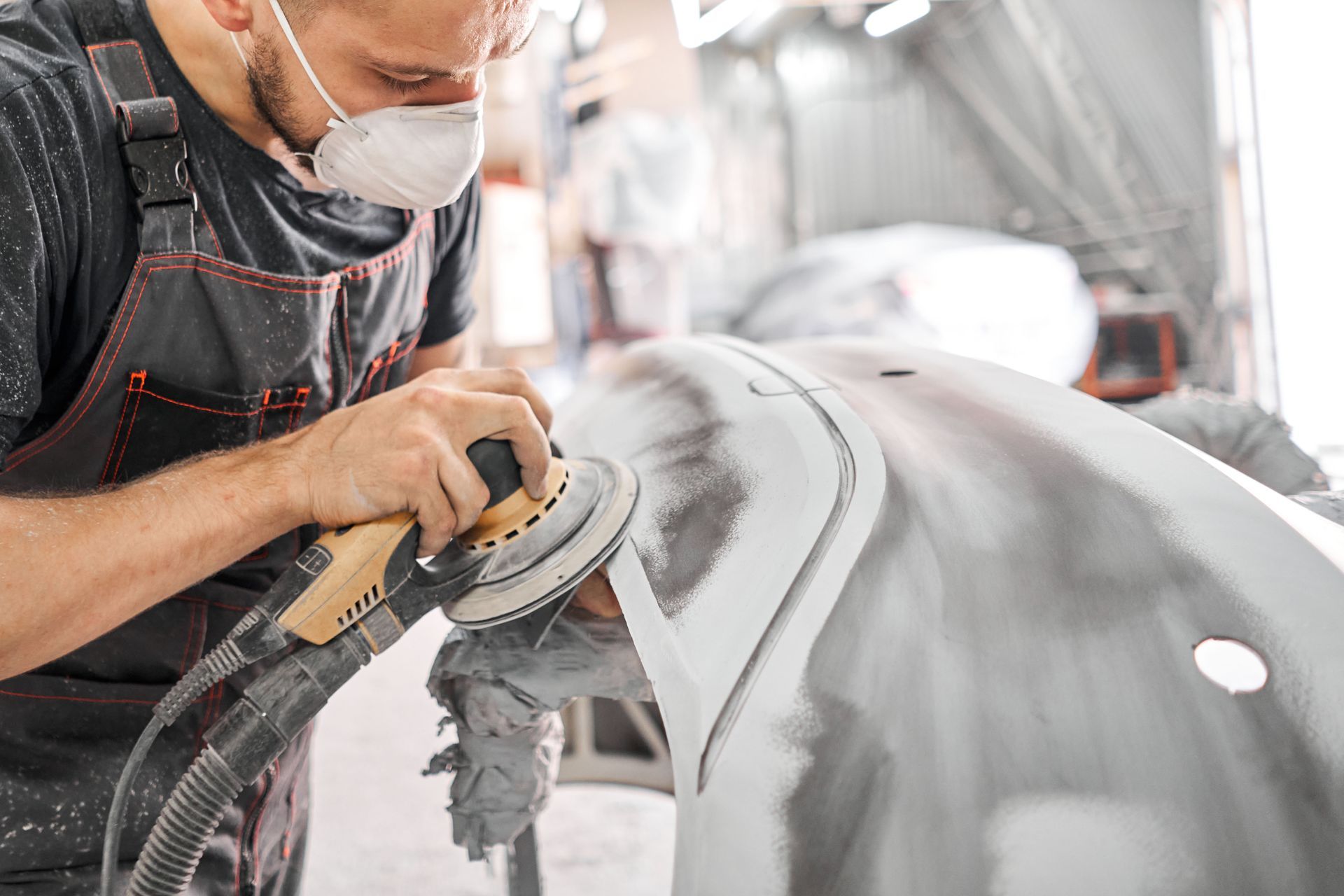 Mechanic sanding car body, wearing mask in a repair shop.