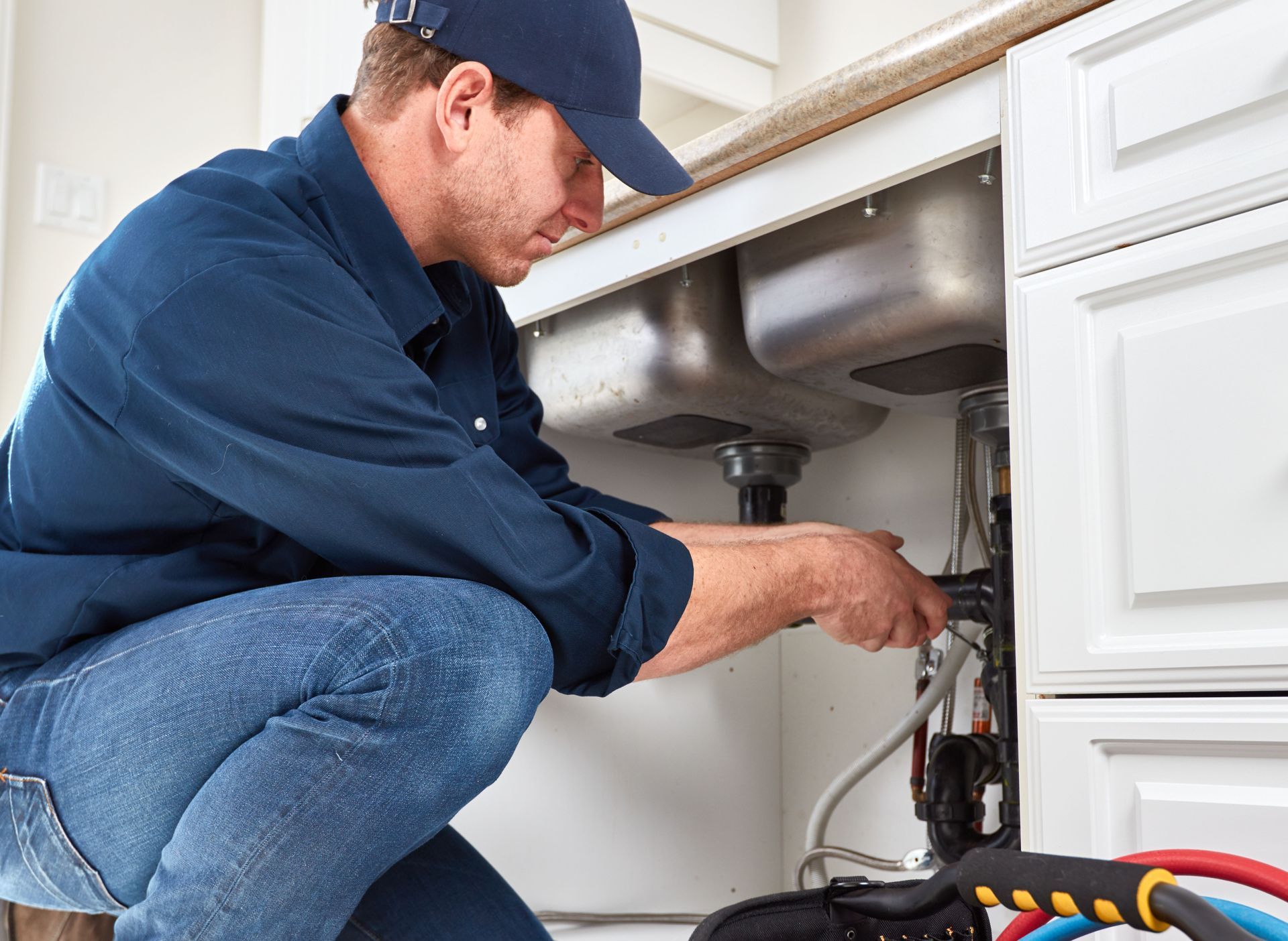 Plumber fixing pipes under a kitchen sink. Wearing blue work shirt, jeans, and cap.