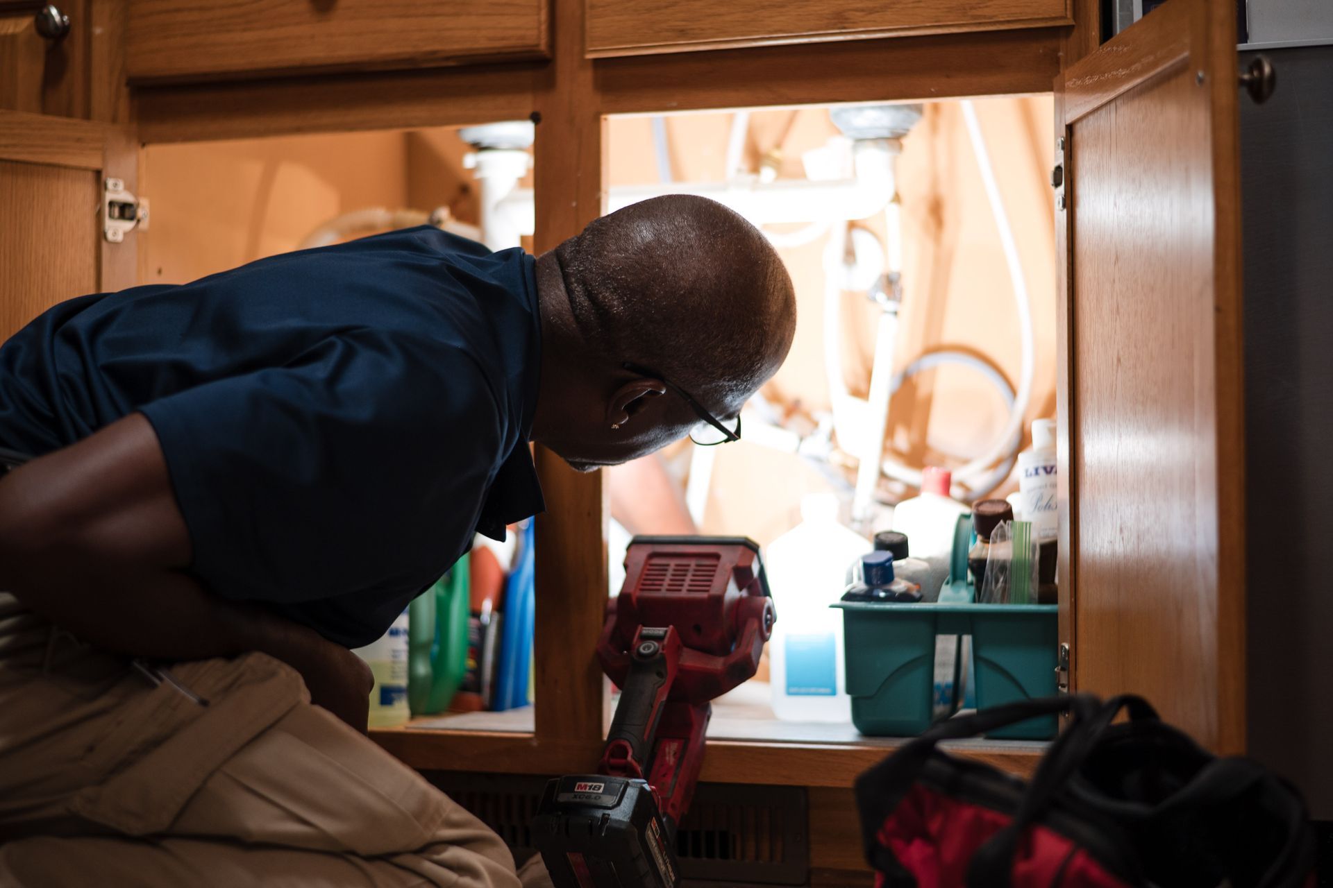 Person kneeling in a wooden cabinet, peering into a bright open appliance area with tools nearby