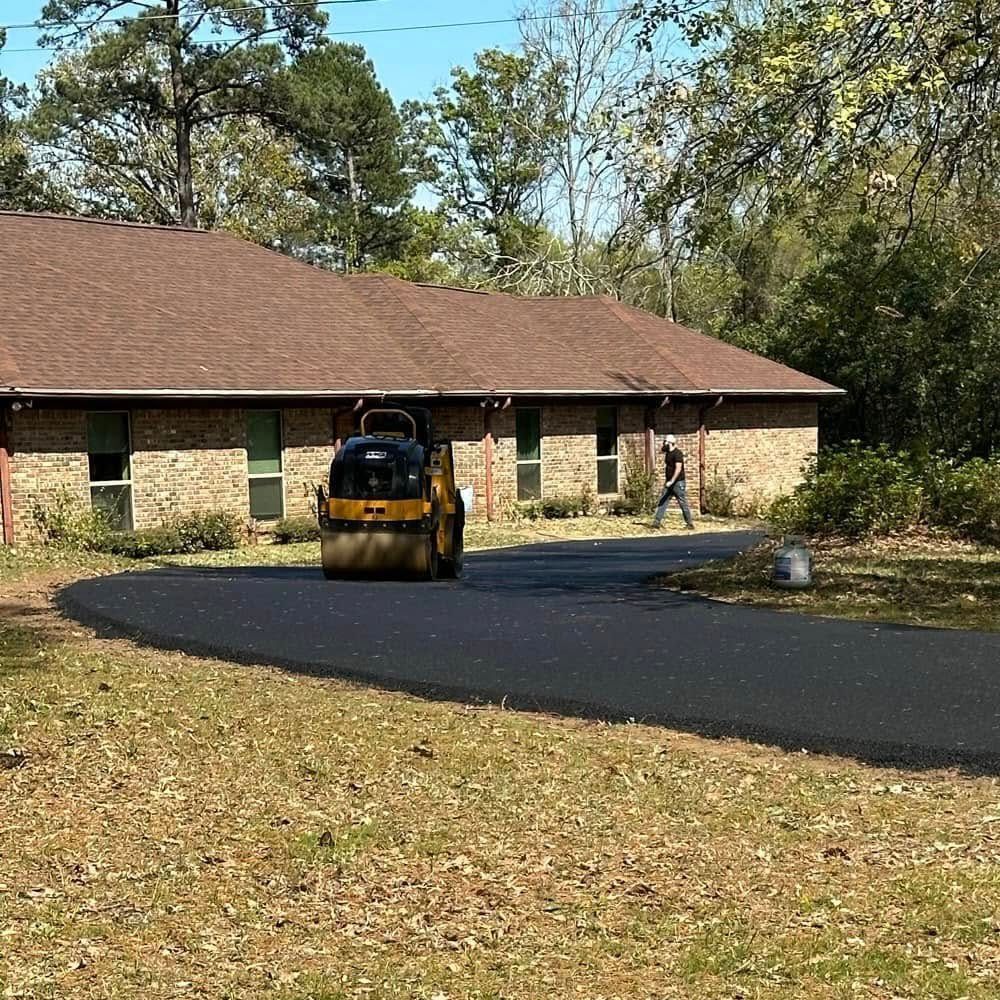 A small yellow road roller smoothing fresh black asphalt on a driveway in front of a stone house with a brown roof.