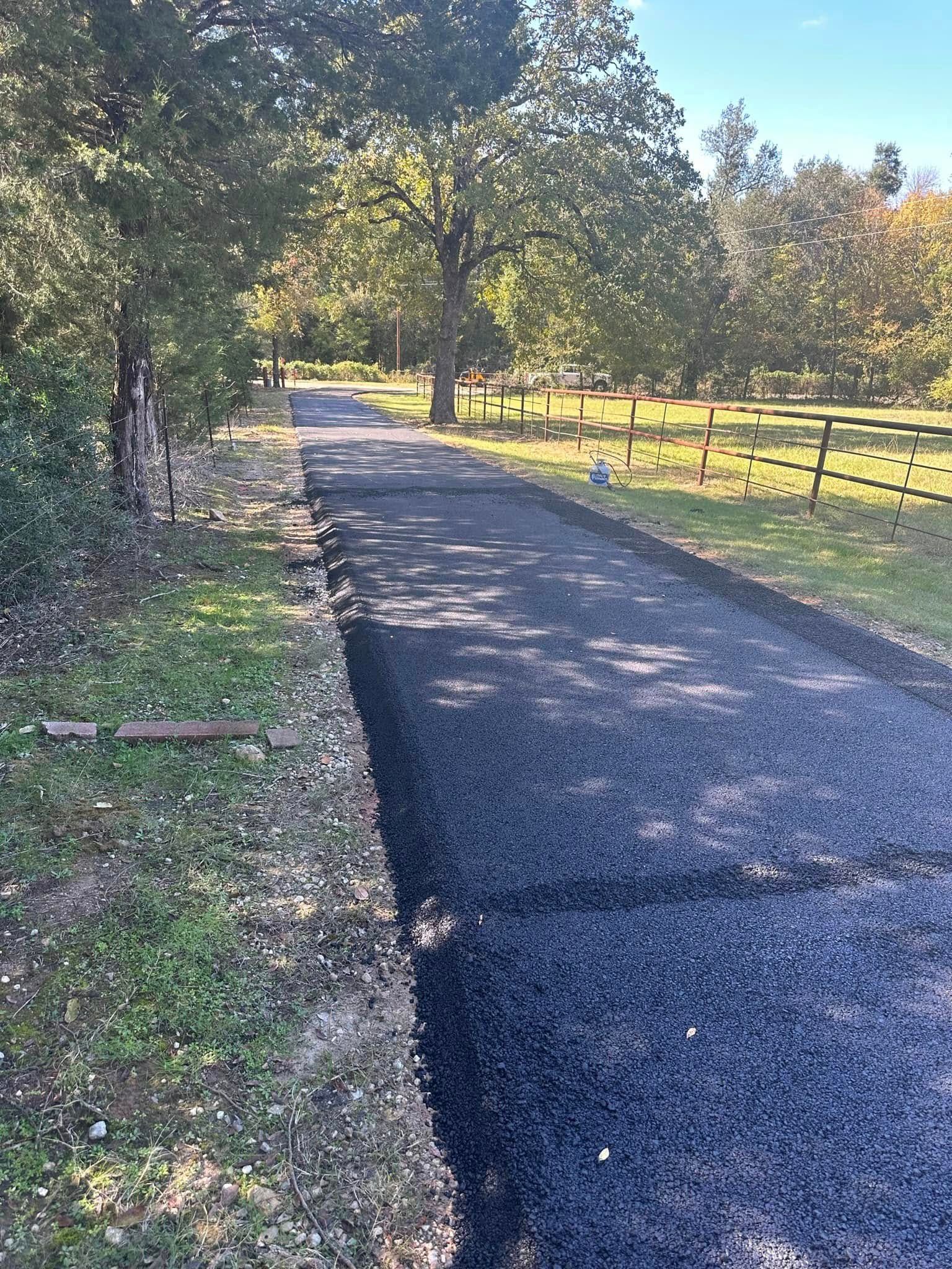 A freshly paved asphalt driveway extends through a tree-lined yard with a wooden fence on the right.