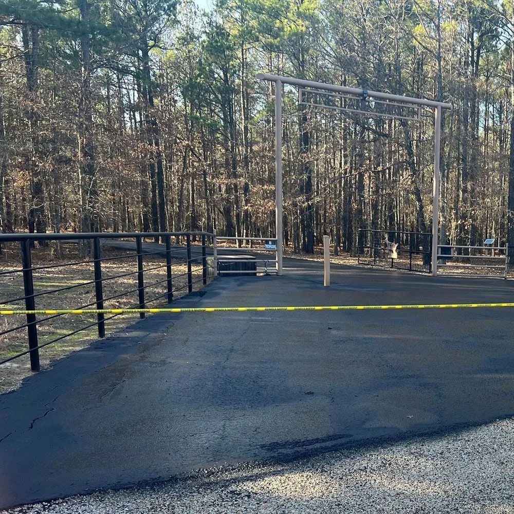 A newly paved black asphalt driveway transitions into a gravel area, framed by black metal fencing and a forest background.