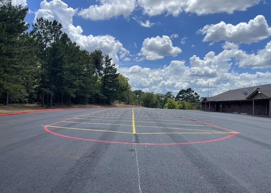 A paved lot featuring painted red and yellow boundary lines on the asphalt, with a treeline and a building in the back.