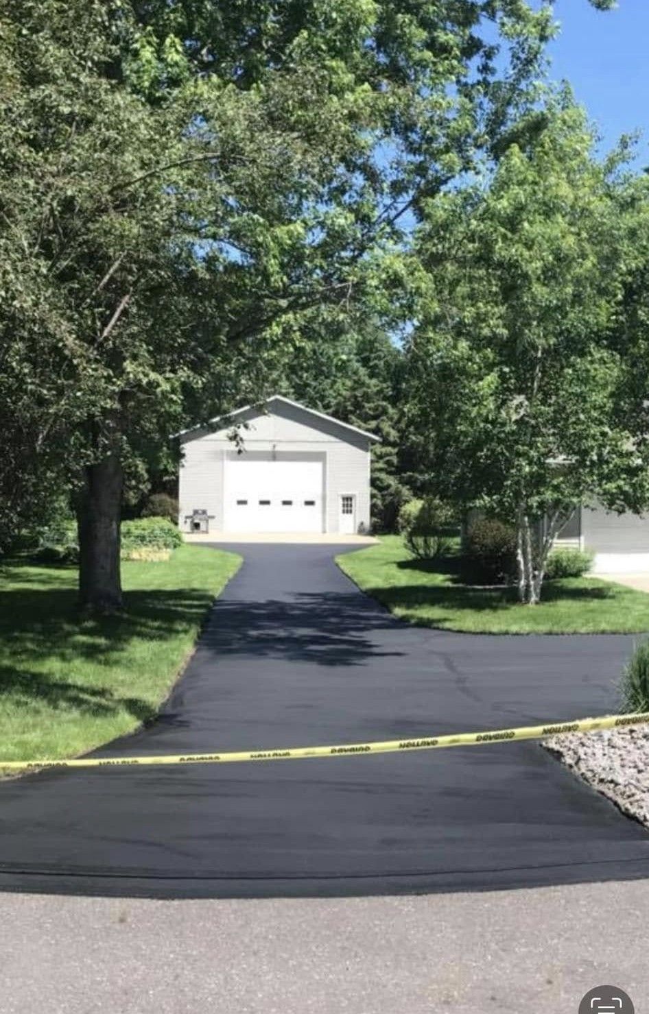 A freshly paved black asphalt driveway leads to a white garage, blocked by yellow caution tape.
