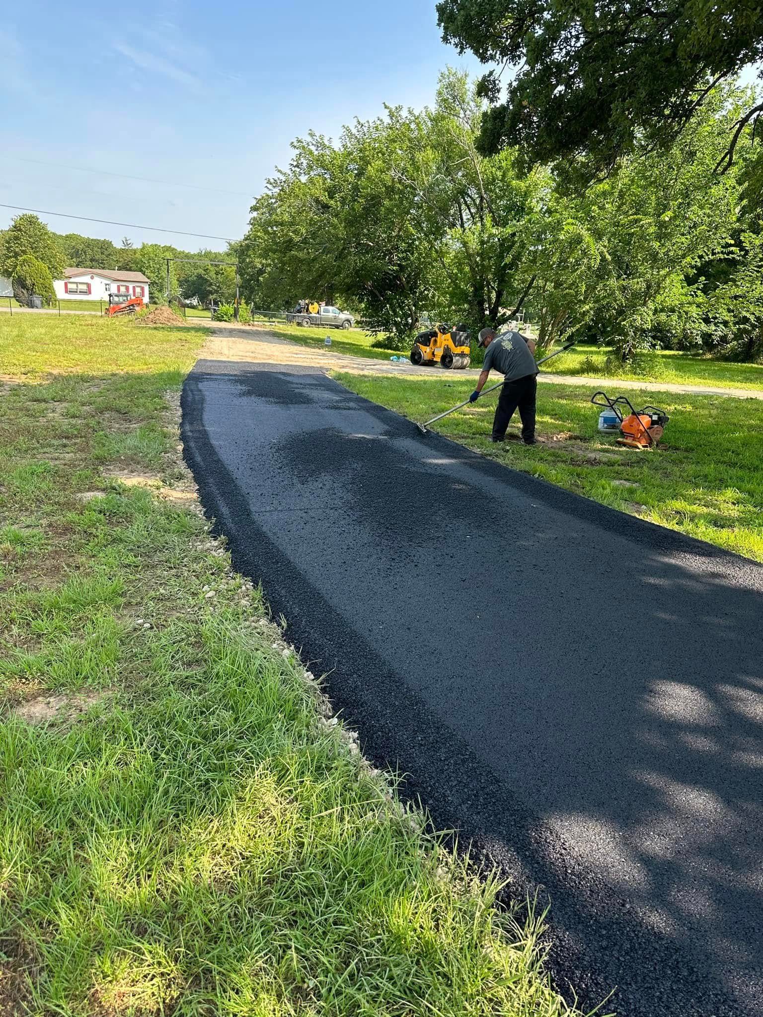 A person spreads loose dark gravel on a newly laid driveway surface next to a grassy lawn under a sunny sky.