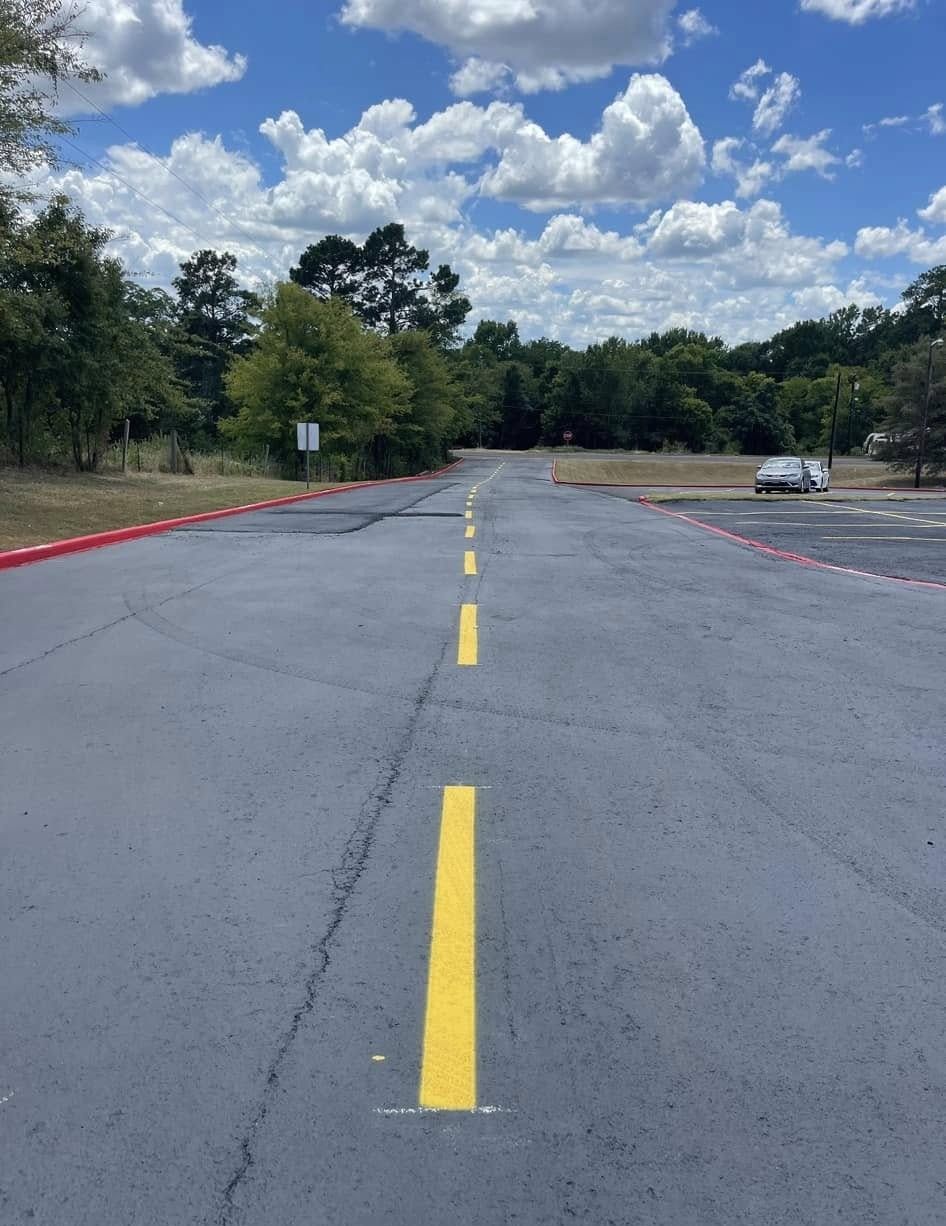 A paved road with a yellow dashed centerline, flanked by red-painted curbs, leading toward trees under a cloudy blue sky.