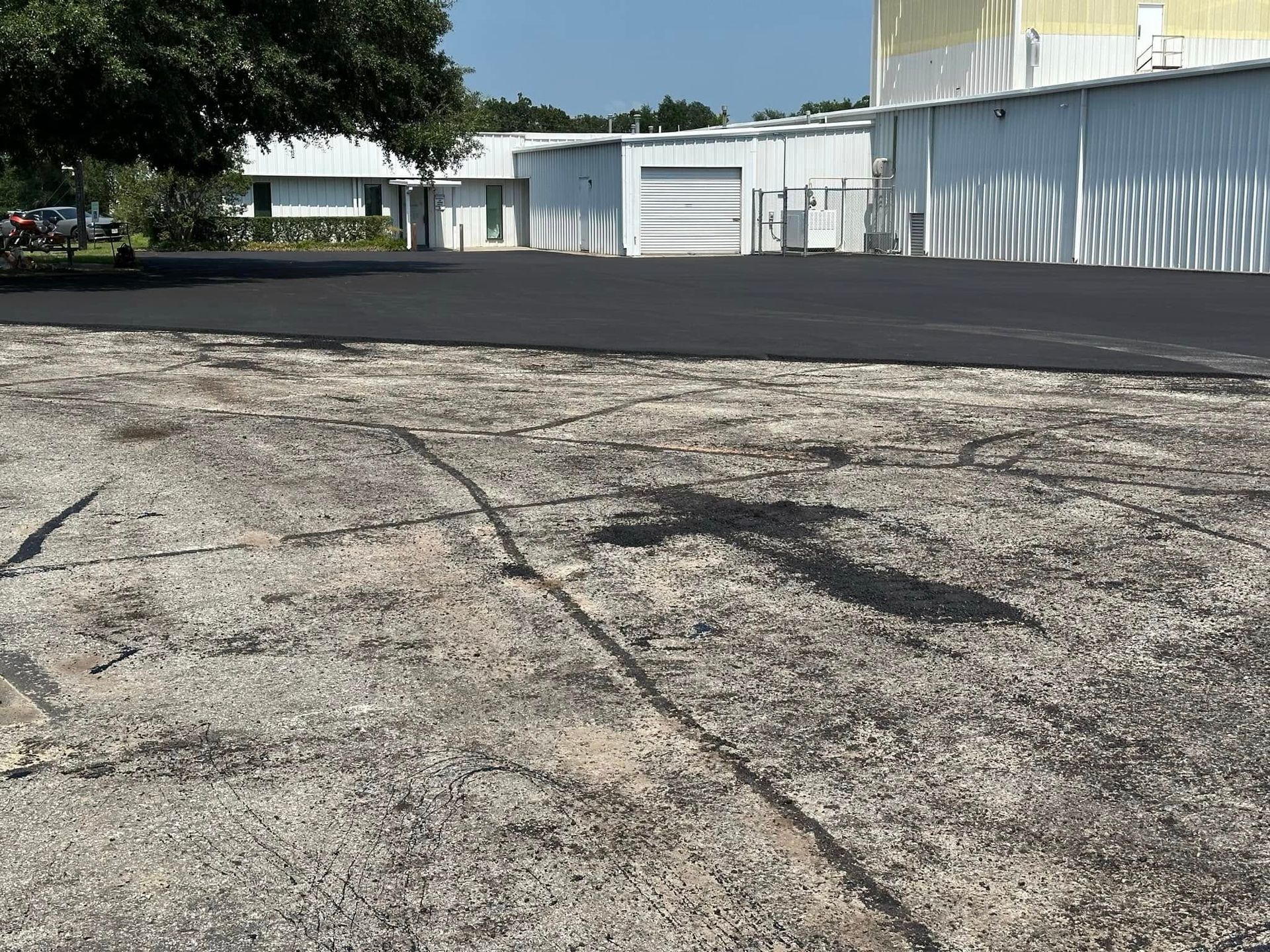 A section of old, cracked gravel paving transitioning to a smooth, newly laid black asphalt surface next to white buildings.