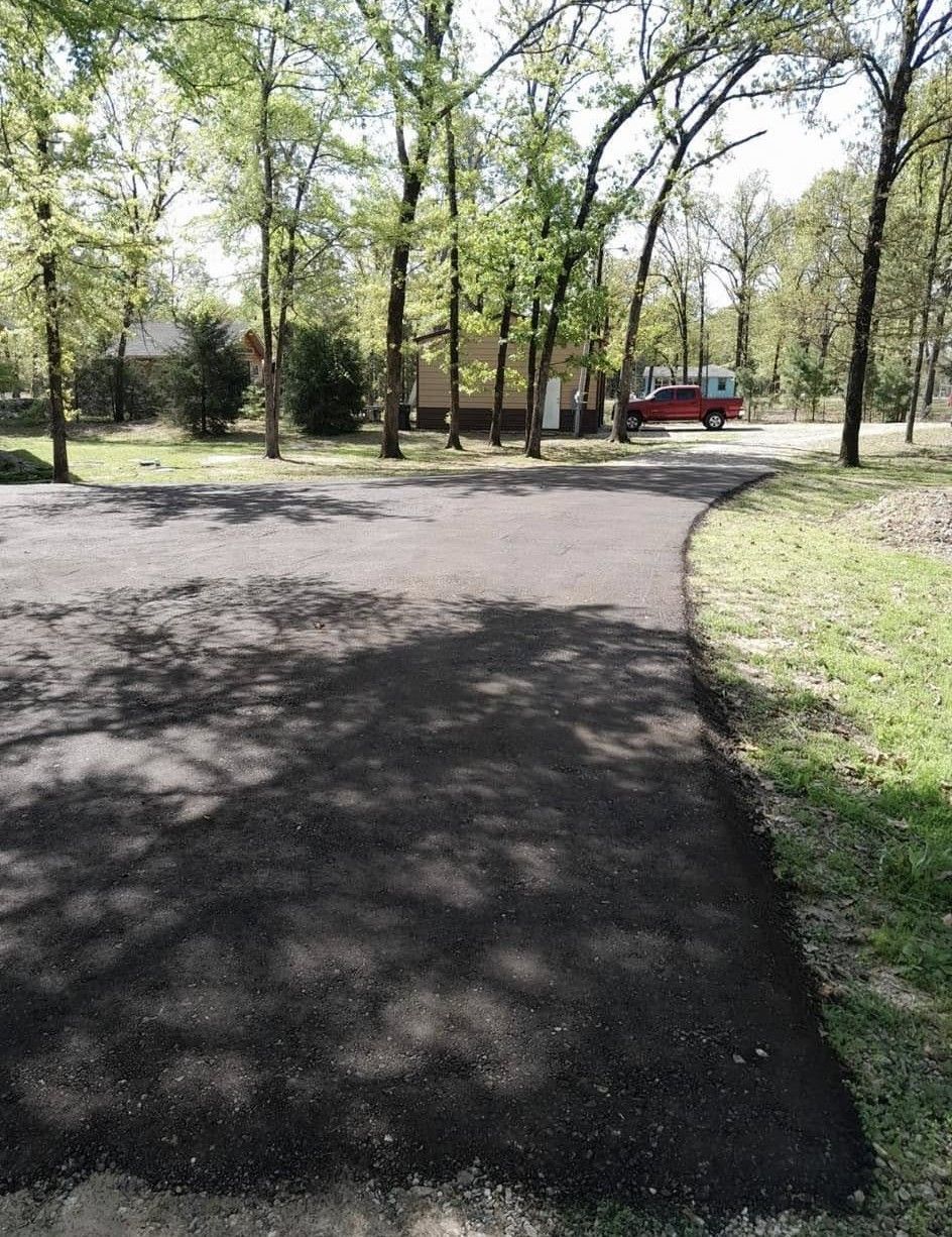 A freshly paved dark asphalt driveway curves through a wooded residential area, leading toward a house and a red truck.