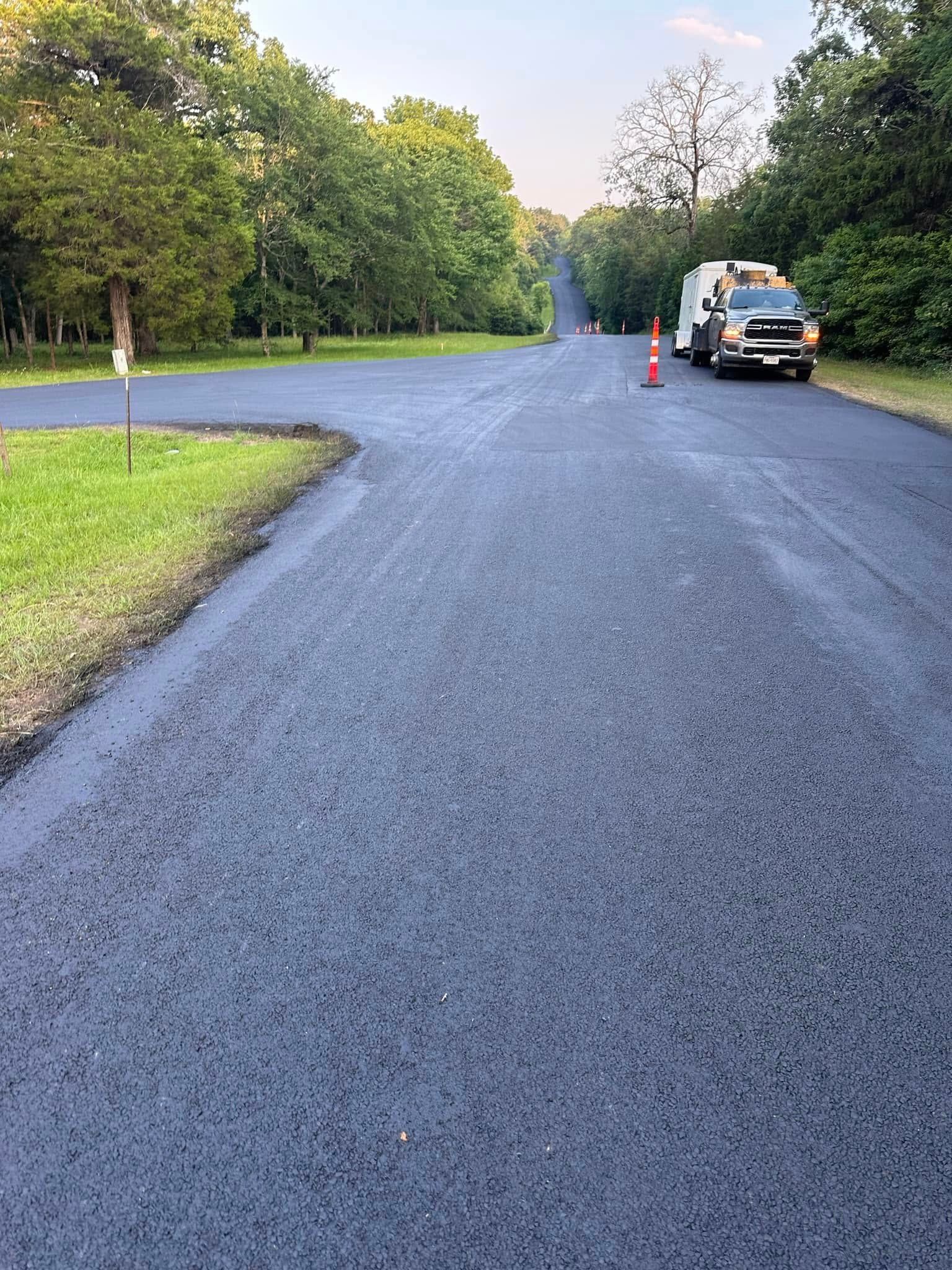 A freshly paved asphalt road curves through a tree-lined area, with a construction truck parked on the side.