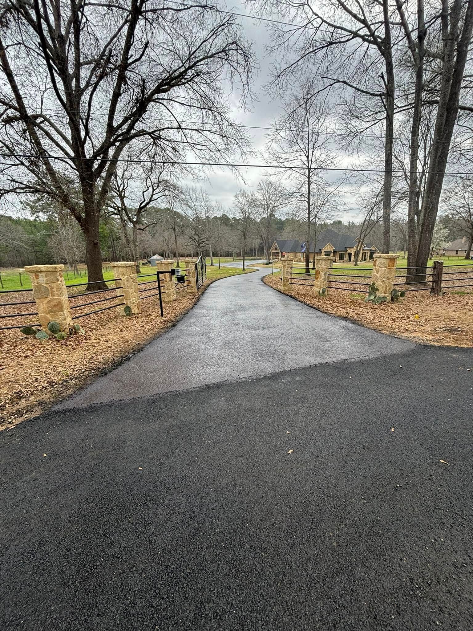 A freshly paved asphalt driveway leading through a rural landscape lined with trees and stone pillars.
