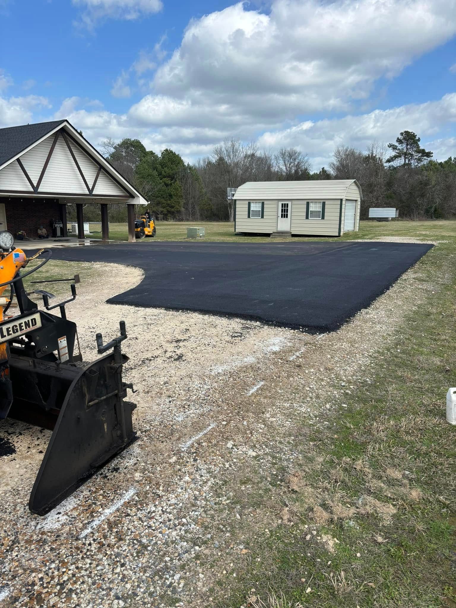 A newly paved asphalt driveway sits in front of a small house with a matching light-colored shed in a grassy field.