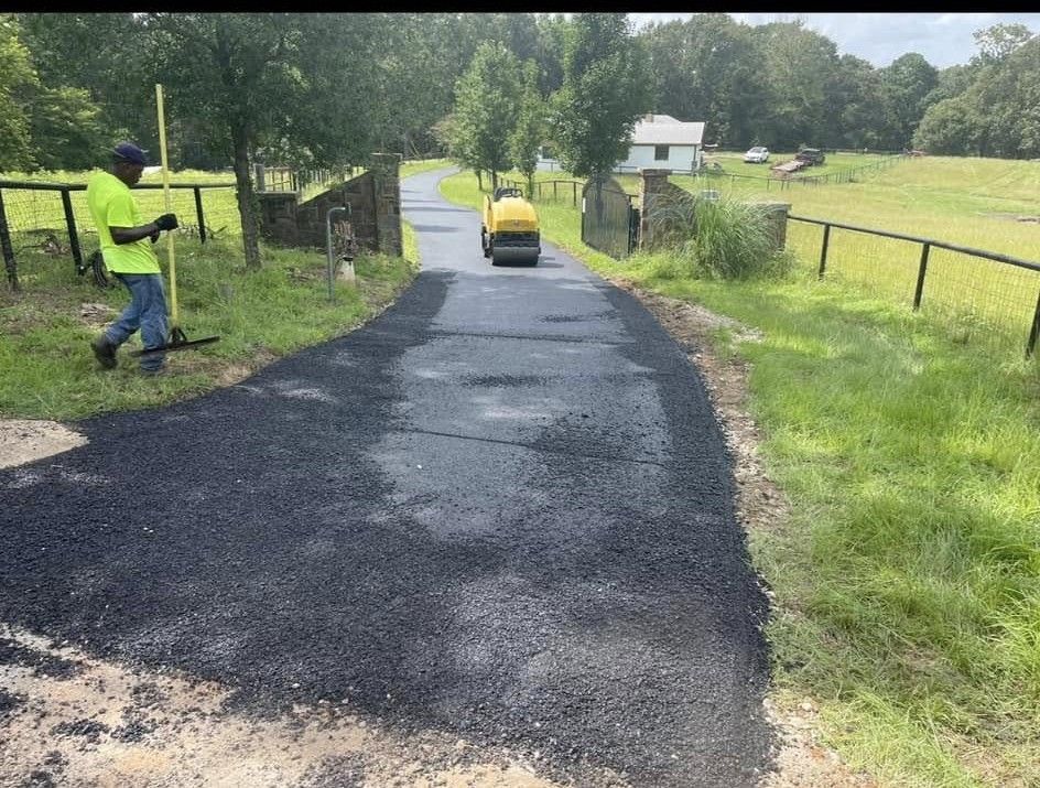 A construction worker in a high-visibility vest stands by a freshly paved driveway, while a steamroller works ahead.