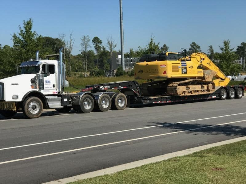 A white semi truck is carrying a yellow excavator on a trailer.