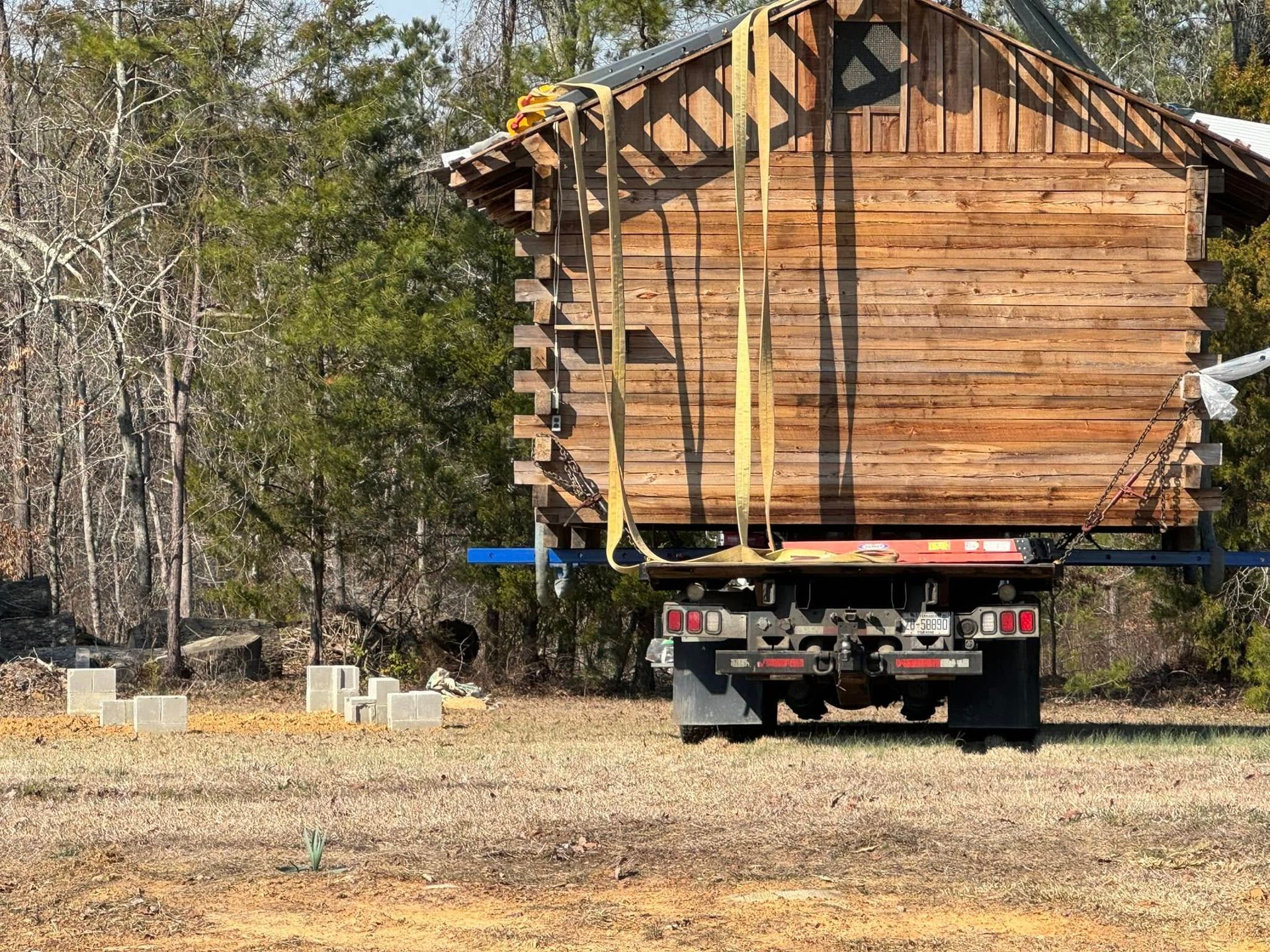 A truck is carrying a log cabin in a field.