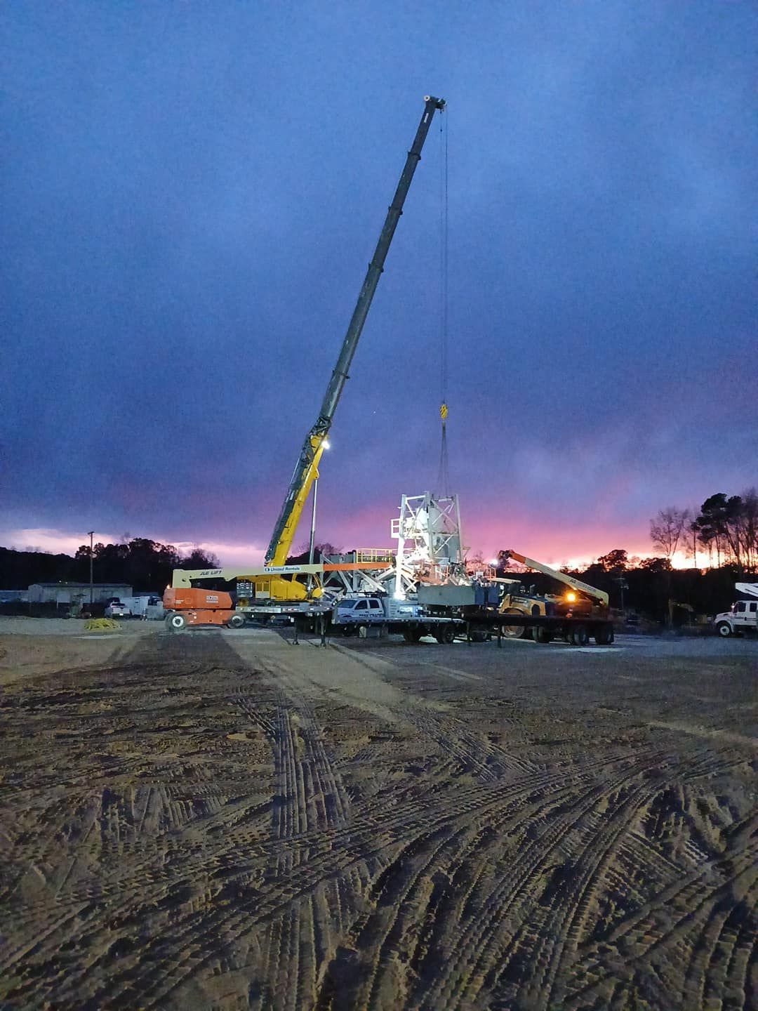 A large crane is lifting a large object in a dirt field at sunset.