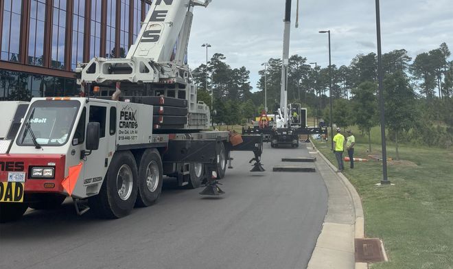 Two large cranes on a road near a building, workers in vests.