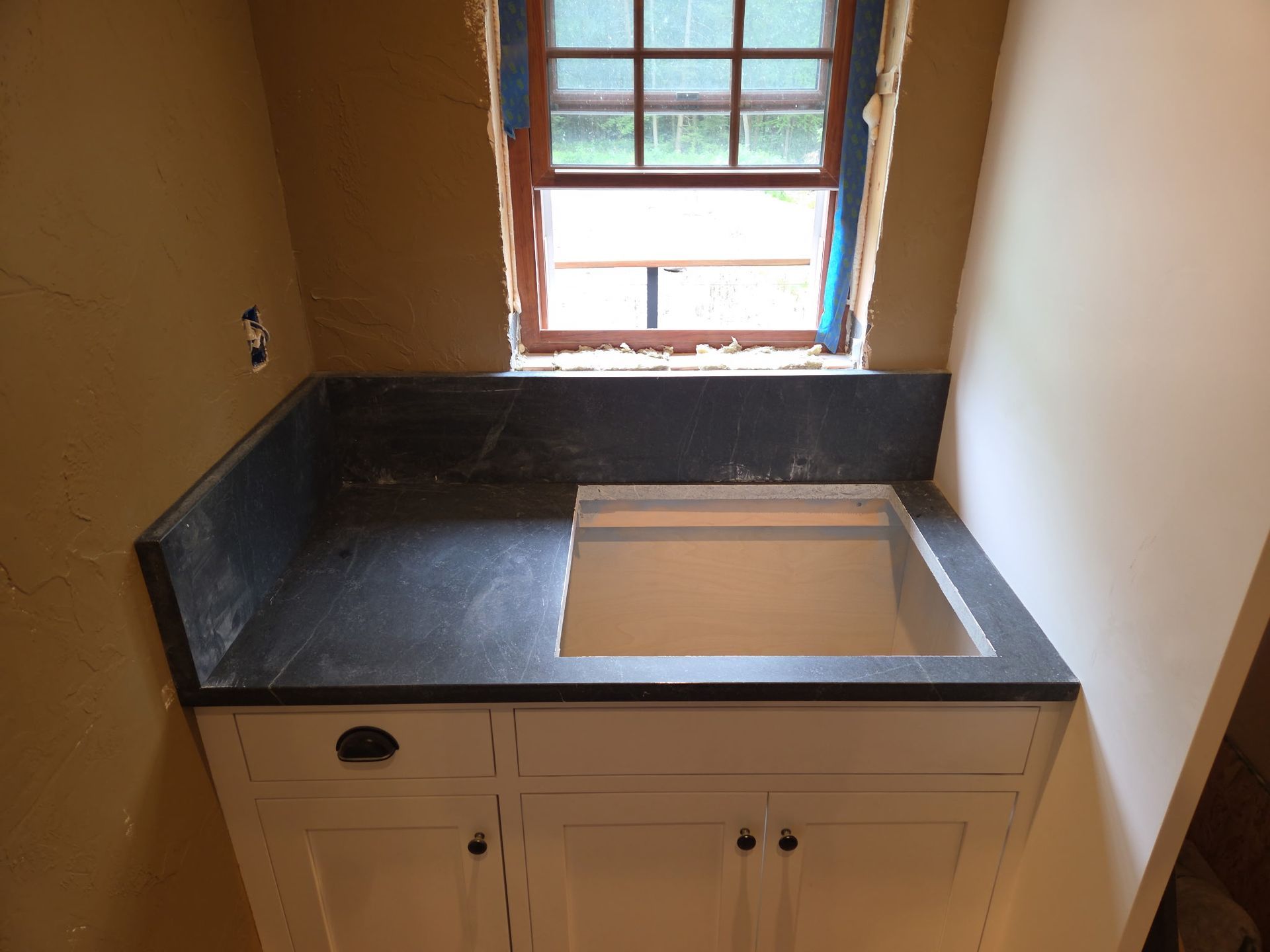 Small kitchen sink with dark countertop beneath a window, surrounded by unfinished walls.