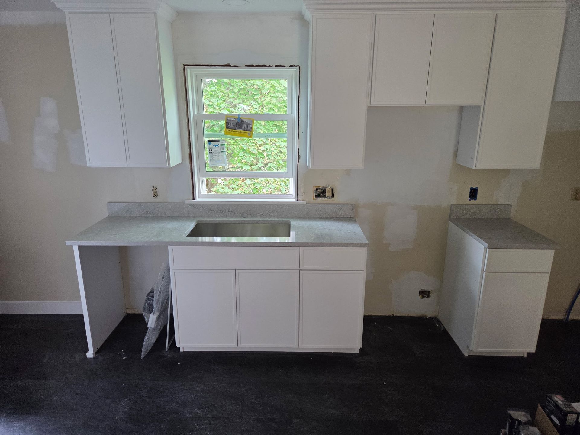 White kitchen cabinets and gray countertops around a window in a partially renovated room.