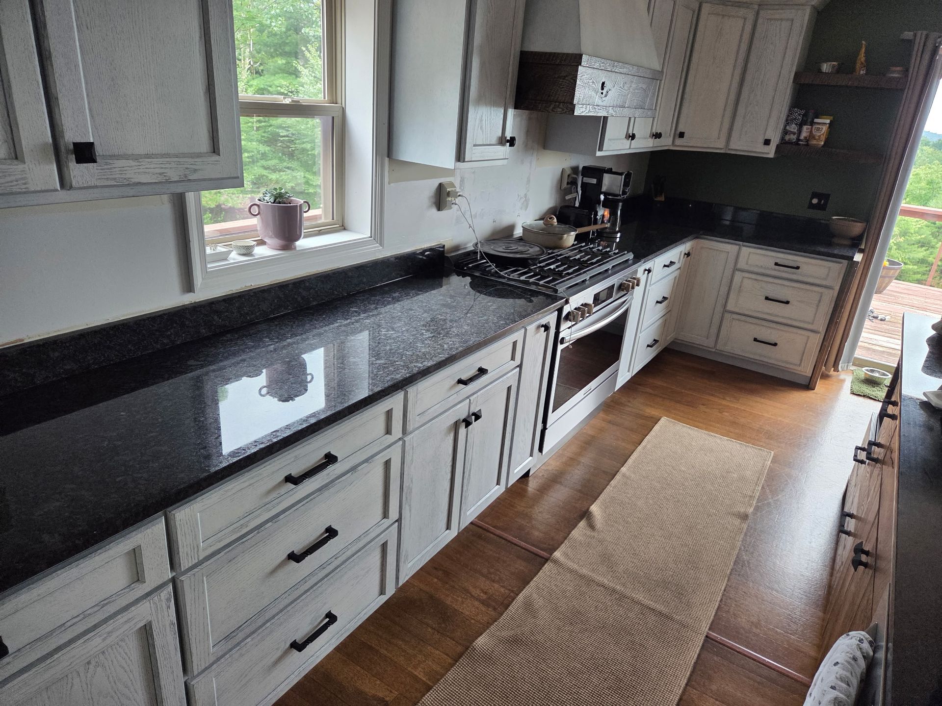 Bright kitchen with white cabinets, black countertop, stainless stove, and a wooden floor runner by the sink.