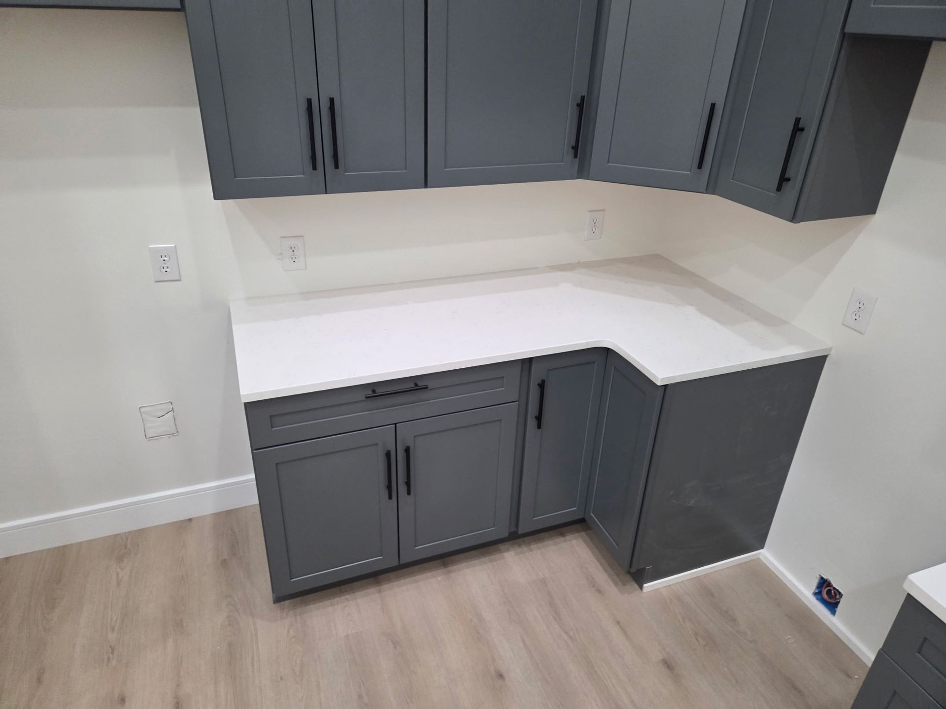 Gray cabinets and white countertop in an empty corner home office nook with wood flooring.
