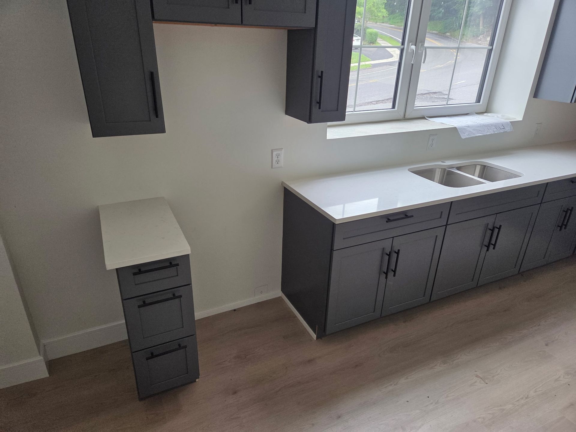 Empty modern kitchen with dark cabinets, white countertops, sink, and window.