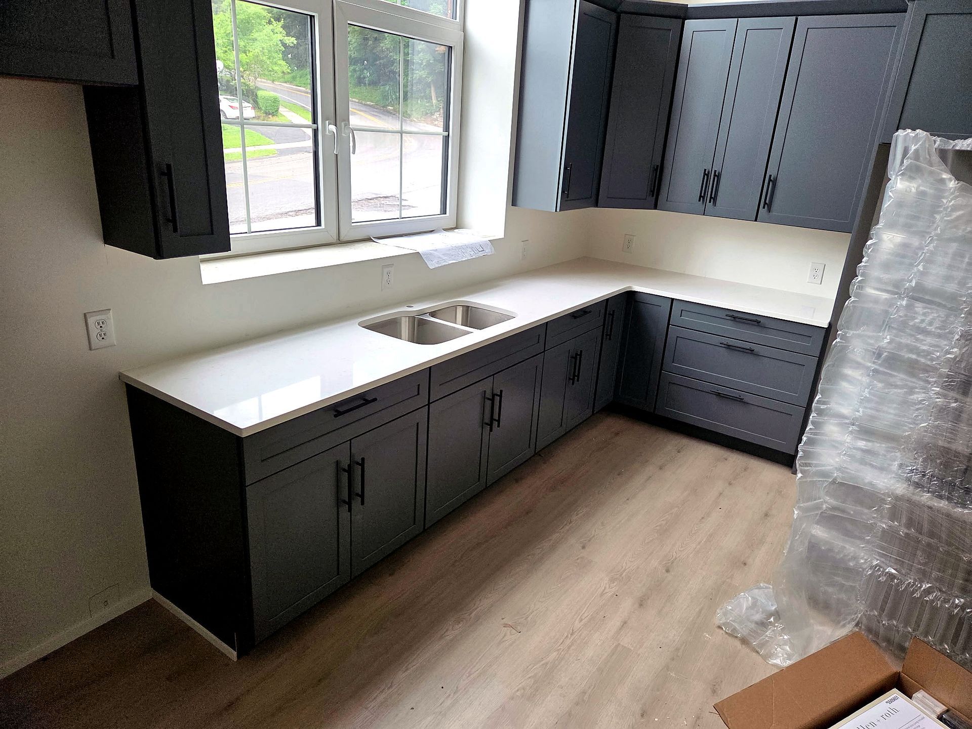 Empty kitchen with dark cabinets, white countertop, window over sink, and light wood flooring.