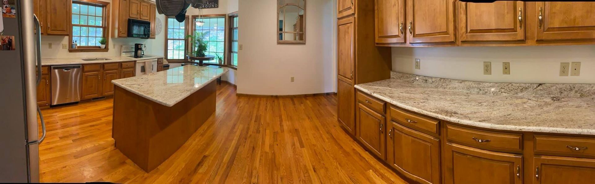 A kitchen with wooden cabinets, granite countertops, an island, and wood flooring. Natural light streams in.