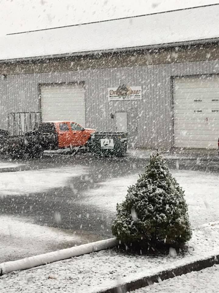 Snow falling on a building with two garage doors and an orange truck parked outside.