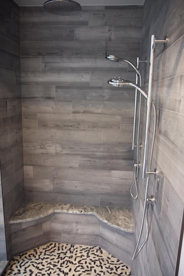 Shower stall with gray wood-look tile, granite bench, and multiple shower heads; the floor has a patterned mosaic.
