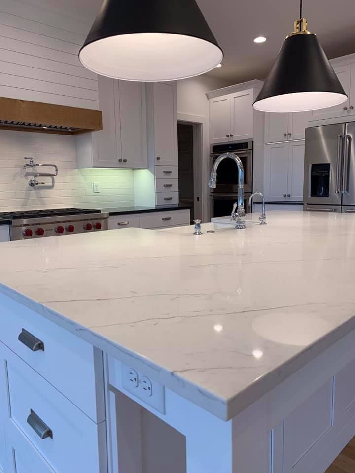 Modern white kitchen with large island, marble countertop, black pendant lights, and stainless steel appliances.