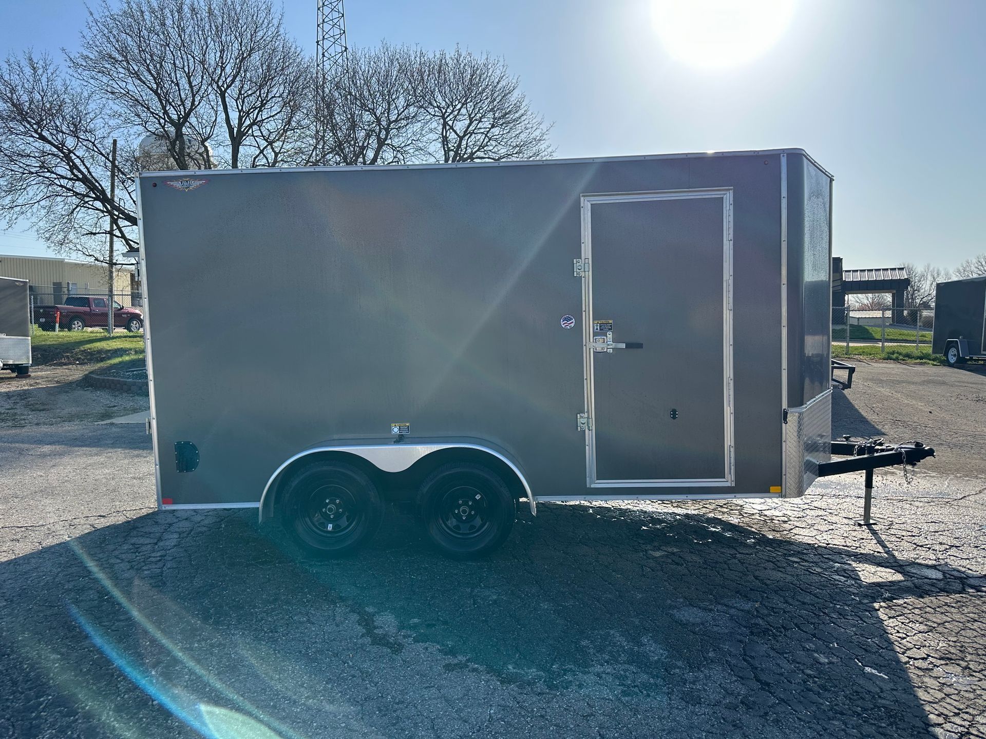 Gray enclosed cargo trailer on gravel with black tires and a door. Sunny outdoor setting.