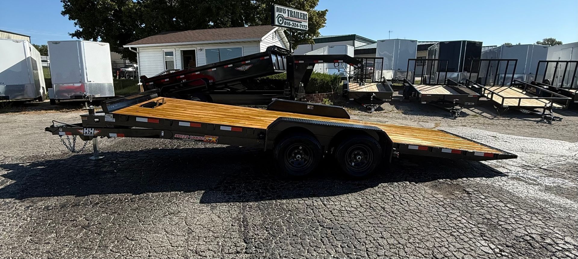 A large, wooden-decked trailer parked on gravel, with other trailers and a building in the background