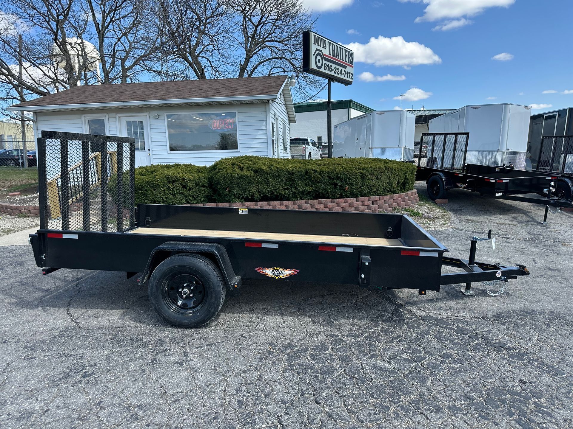 Black utility trailer with black railings and a wood bed on a paved surface near a building.
