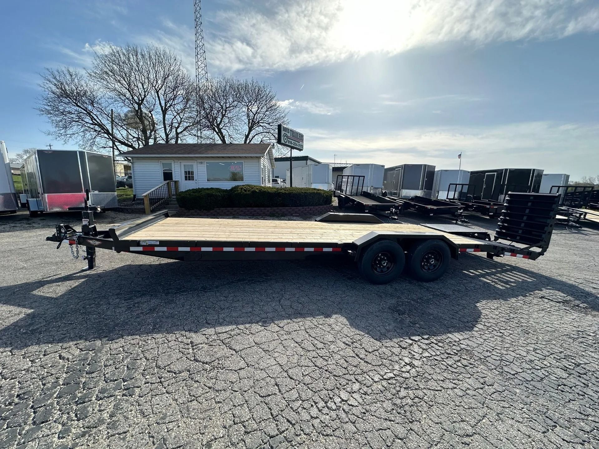 Black flatbed trailer parked on gravel, with a building and other trailers in the background. Sunny day.